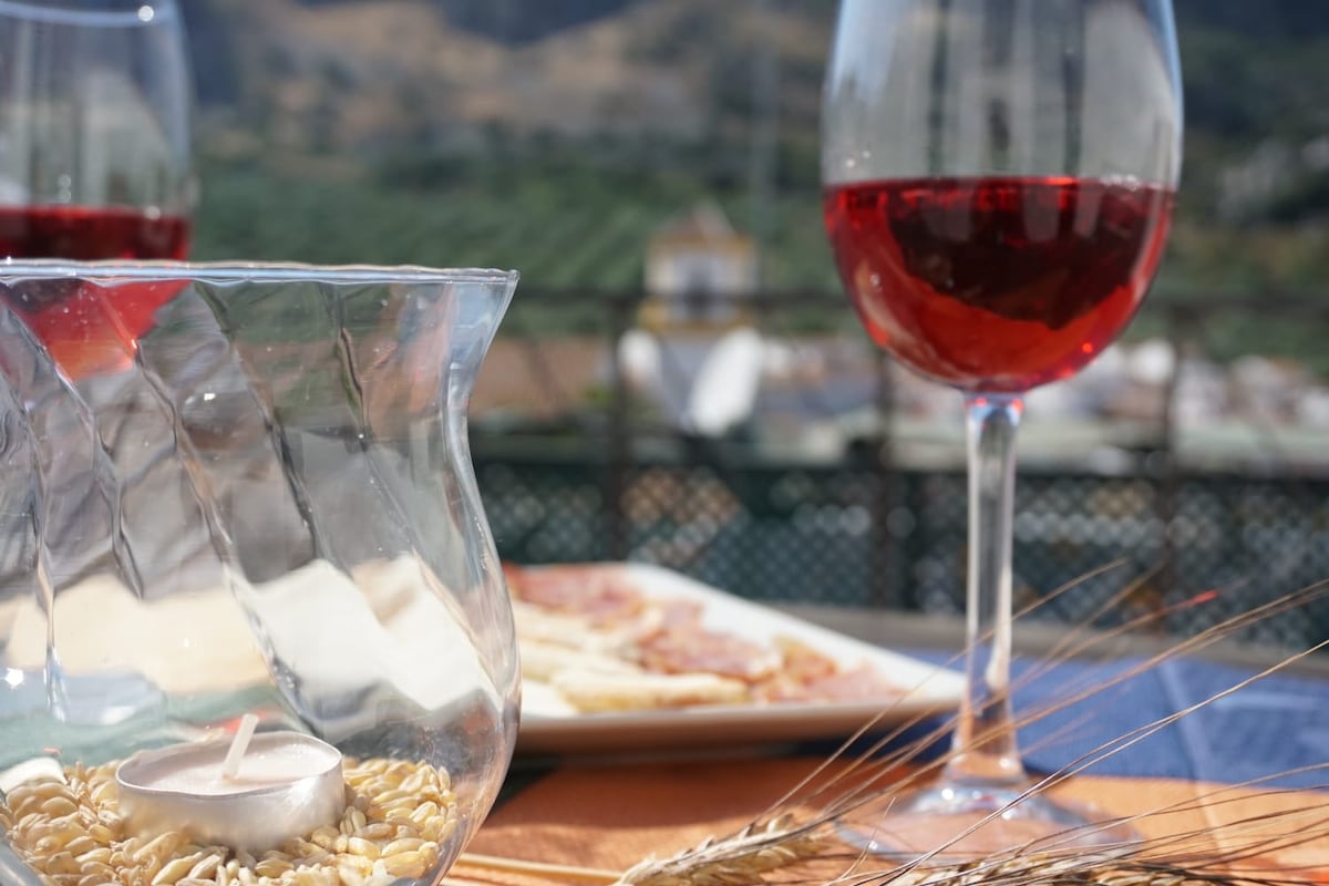 A table setting features a glass of red wine beside a plate of sliced meats. A candle is placed in a decorative glass holder filled with grains. A scenic backdrop of rolling hills and distant village rooftops is visible.