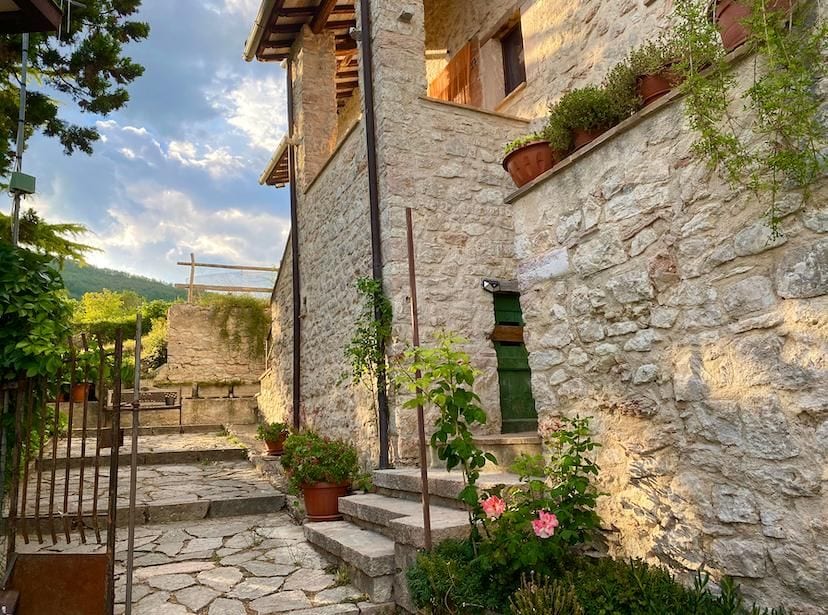 A charming stone pathway leads through a landscaped area, framed by planters with green foliage and flowers. The rustic stone walls of the building create a warm backdrop, while the soft evening light enhances the serene atmosphere of the outdoor space.