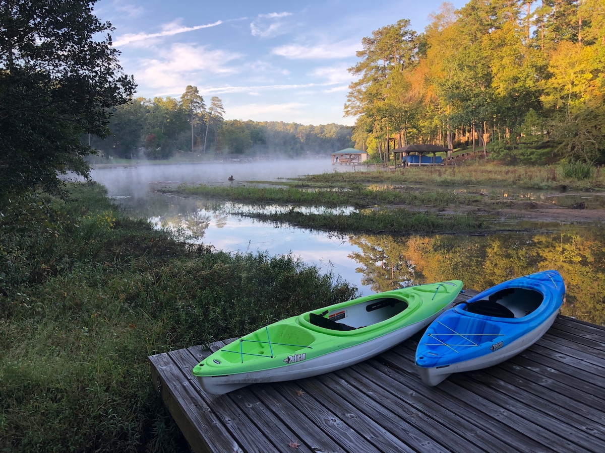 Two kayaks sit on a wooden dock overlooking a misty cove. The vibrant green and blue kayaks contrast against the calm water and lush greenery, while trees surround the serene landscape, reflecting a peaceful natural environment.