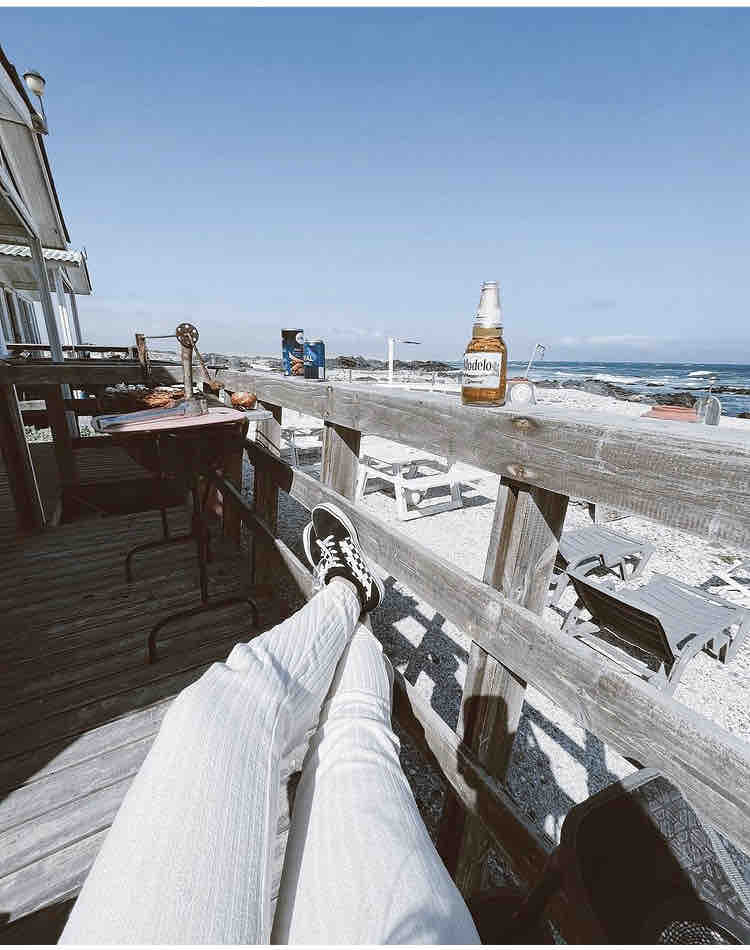 A wooden railing extends along a deck overlooking a pebbled beach with gentle waves. A bottle of beer rests on the railing, accompanied by a plate of food. Tables with chairs are spaced out, inviting relaxation in the sunlit outdoor area.
