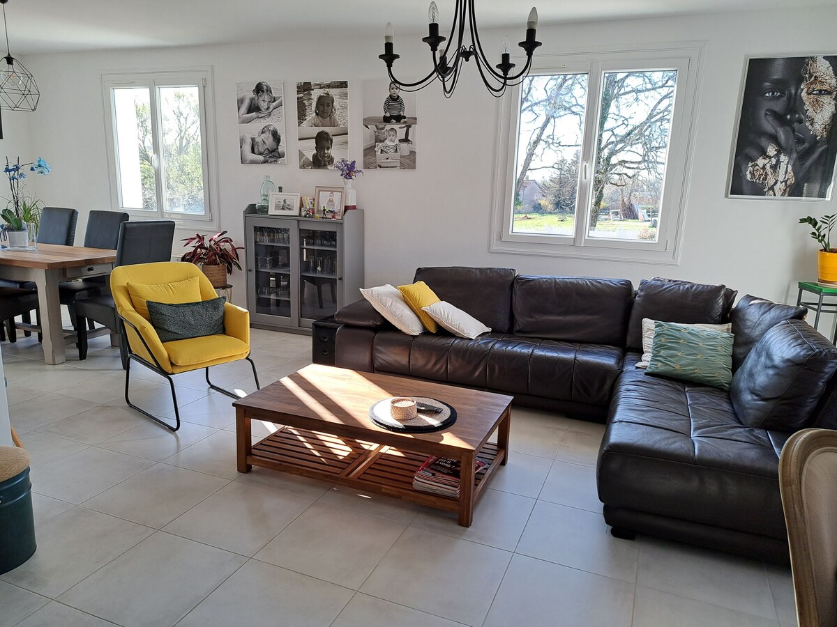 A spacious living area features a dark leather sectional sofa and a wooden coffee table. A bright yellow chair adds a pop of color. Natural light flows through large windows, illuminating the room. A dining table with chairs is visible in the background, accompanied by decorative elements.