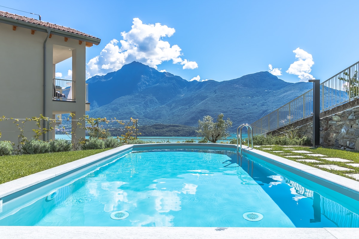 A refreshing swimming pool is visible in the foreground, surrounded by a well-maintained lawn. Landscaped stone pathways lead towards the pool, while panoramic views of the mountains and lake are clearly showcased in the background under a bright blue sky.