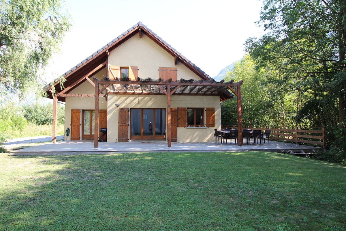 The exterior of a large detached house is depicted, featuring a covered terrace with seating and dining space. Lush greenery surrounds the property, and large glass doors provide access to the living area. The sloped roof and wooden elements contribute to a rustic charm.