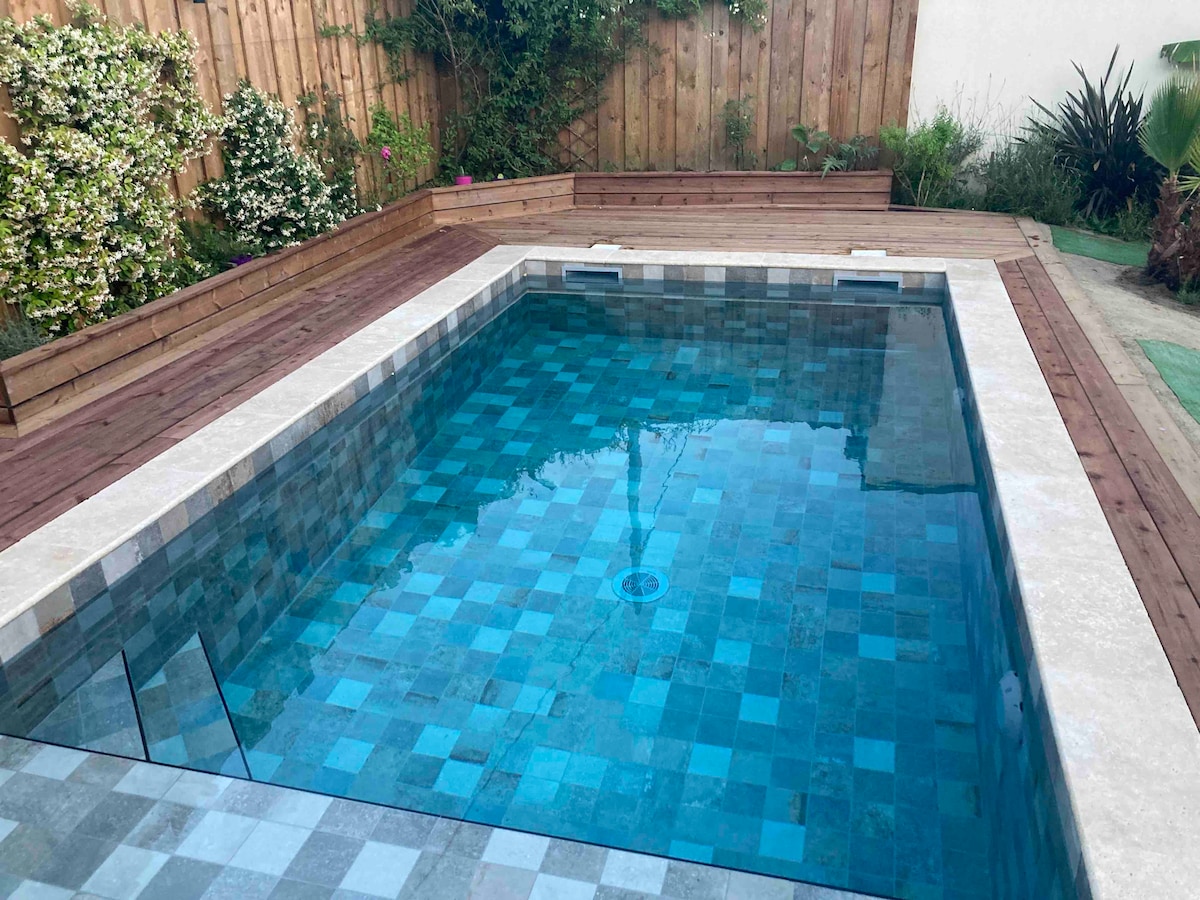 A rectangular pool is set within a wooden deck, surrounded by lush greenery and flowering plants. The pool features a mosaic pattern of blue and gray tiles, reflecting light across the water's surface. The area offers a peaceful atmosphere suitable for relaxation.