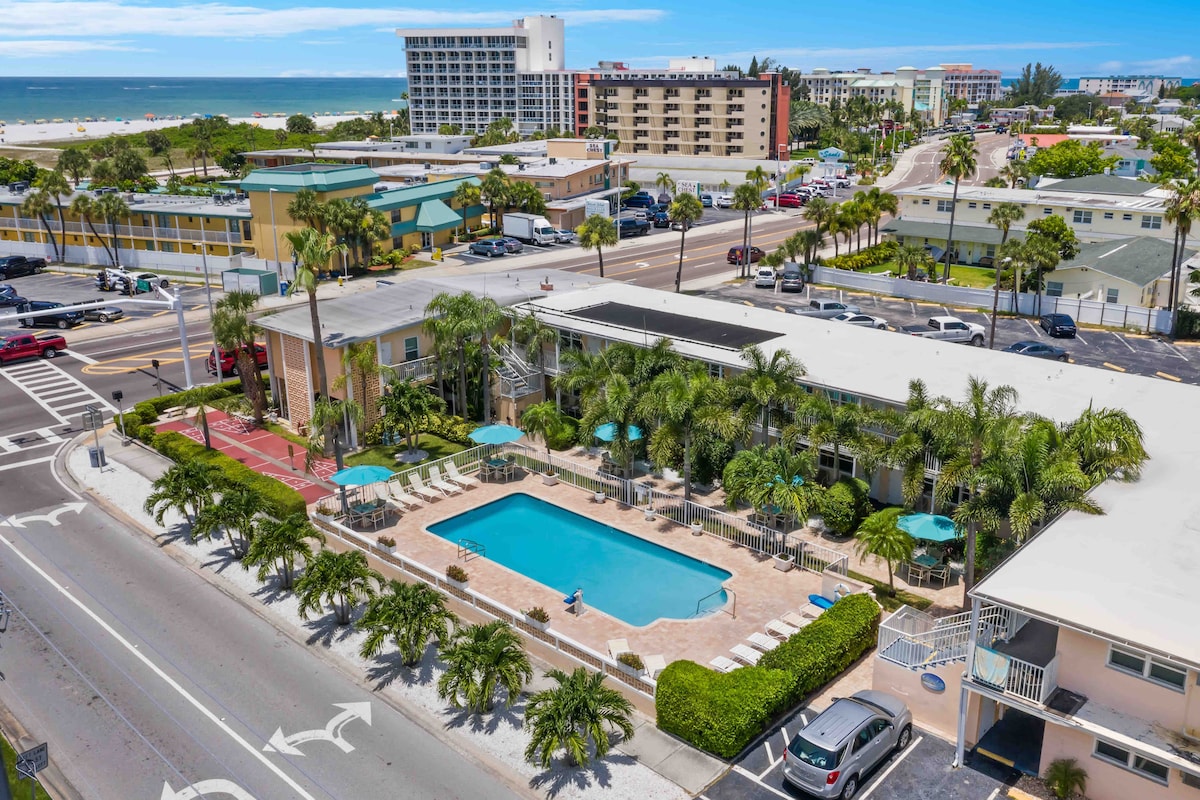 An aerial view captures the vibrant property, featuring a pool surrounded by lounge chairs and palm trees. The beach is visible in the distance, alongside various buildings. The layout suggests easy access for guests and a sunny outdoor atmosphere.