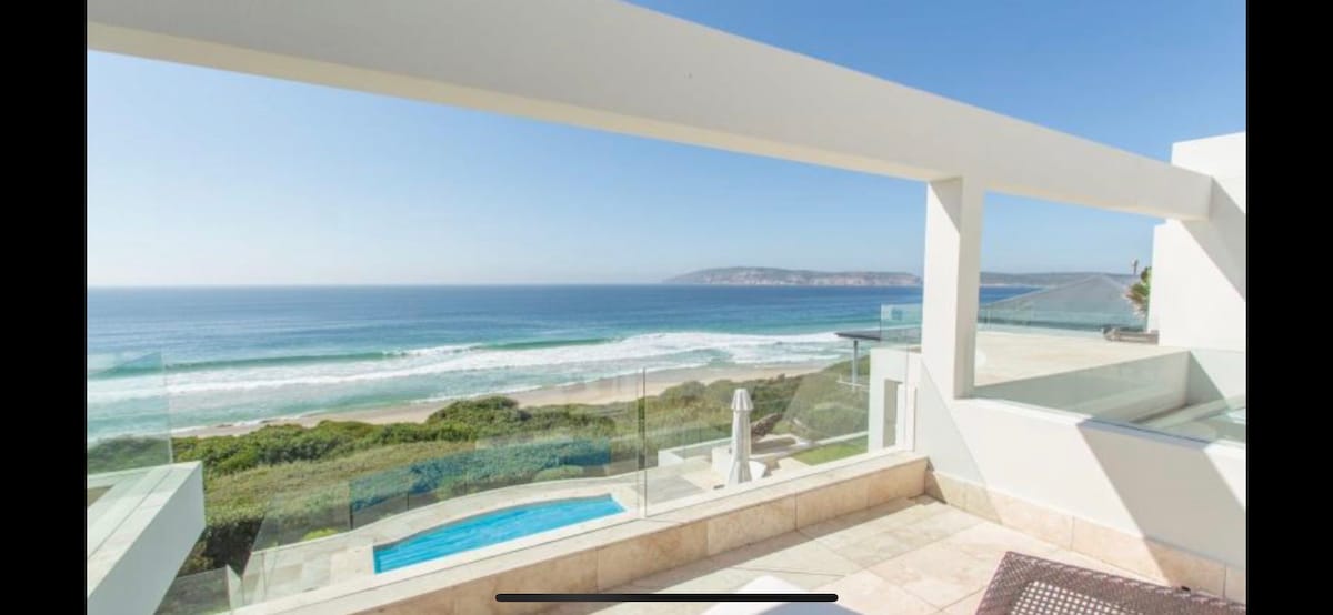 An expansive terrace offers unobstructed views of the coastline, with rolling waves and sandy beach visible below. A sparkling pool is situated adjacent to the deck, framed by modern glass railings. The clear blue sky enhances the serene coastal ambiance.