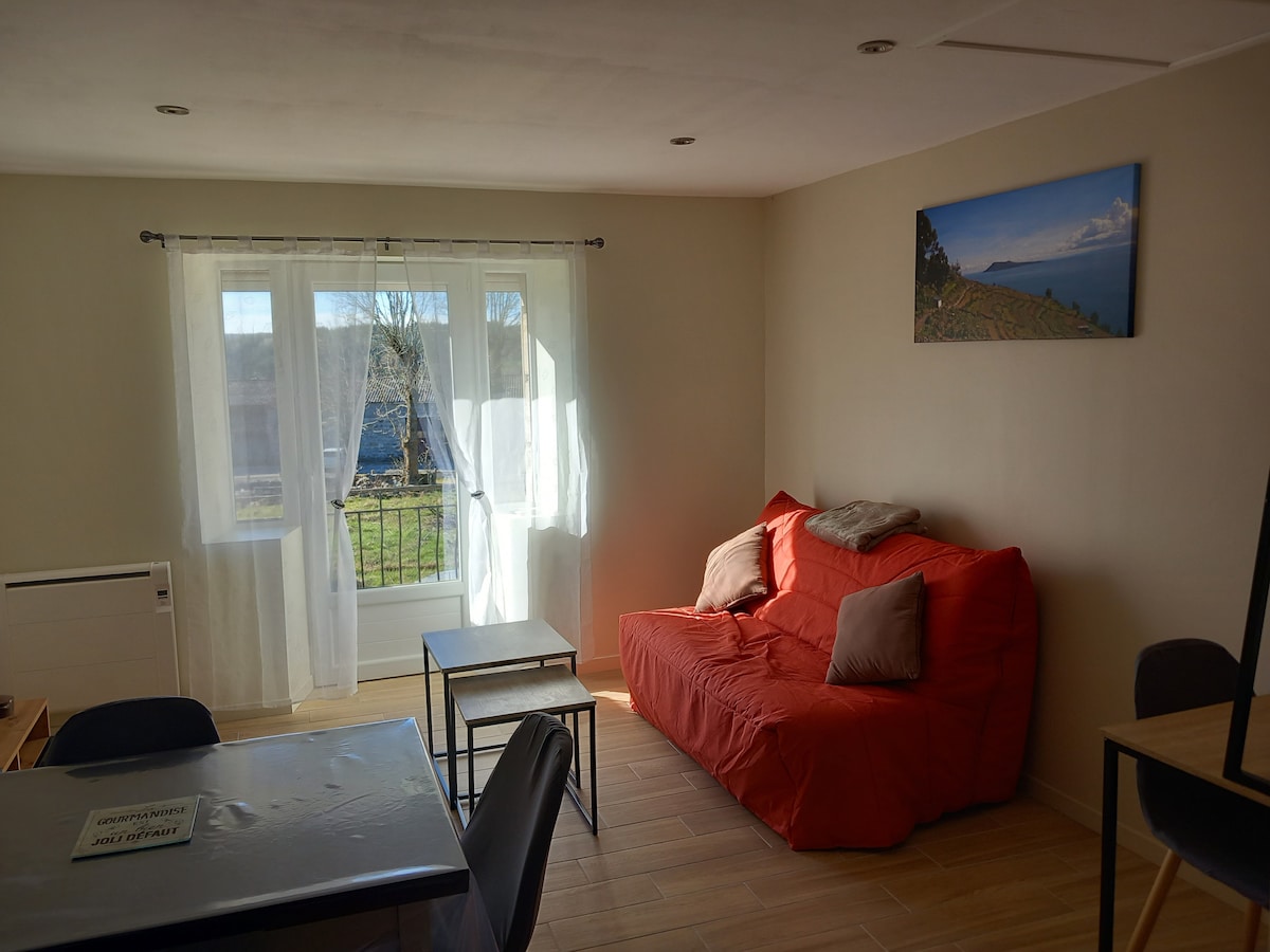 The living area features a red sofa bed complemented by two neutral cushions. A small dining table is positioned nearby, and large windows allow natural light to fill the space. A scenic landscape photograph adorns the wall, enhancing the room's inviting simplicity.