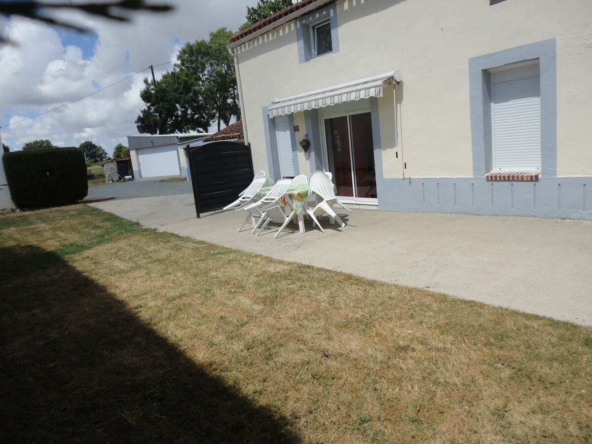 An outdoor patio area is visible, featuring a smooth concrete surface with five white lounge chairs arranged around a small table. A grassy area extends beyond the patio, complemented by shrubbery along the edge. Soft clouds drift in the blue sky above.