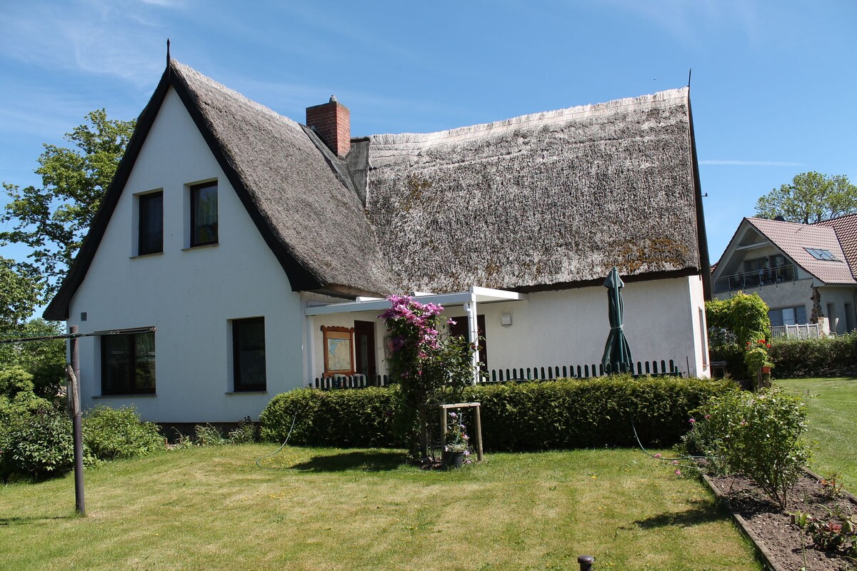 The exterior of a charming thatched-roof house is presented, featuring a well-maintained lawn and flower beds. A patio with umbrellas is visible, surrounded by greenery. The two-story structure displays a white facade, offering a welcoming appearance and a glimpse of the nearby residential area.