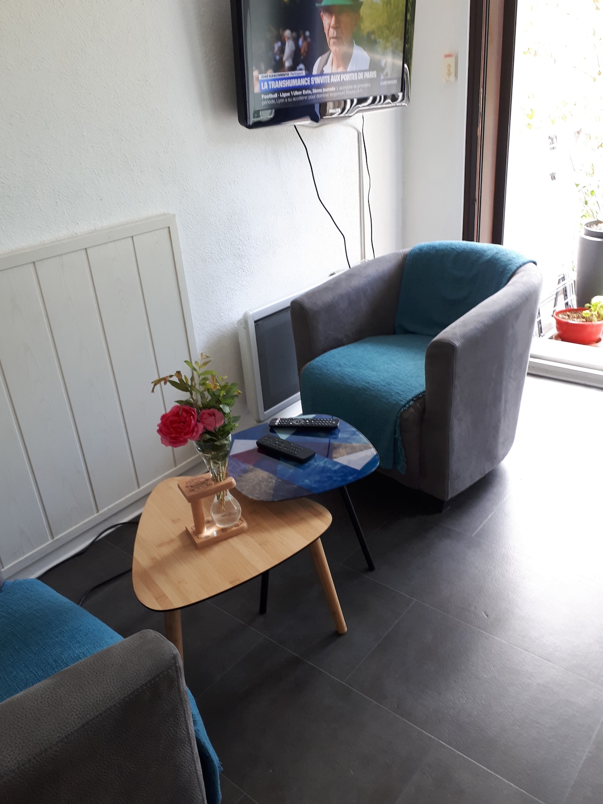 A seating area features two gray and teal armchairs arranged around an angled wooden coffee table. A small vase with flowers sits on the table, and a television mounted on the wall adds functionality to the space. Sunlight is visible through the nearby doors.