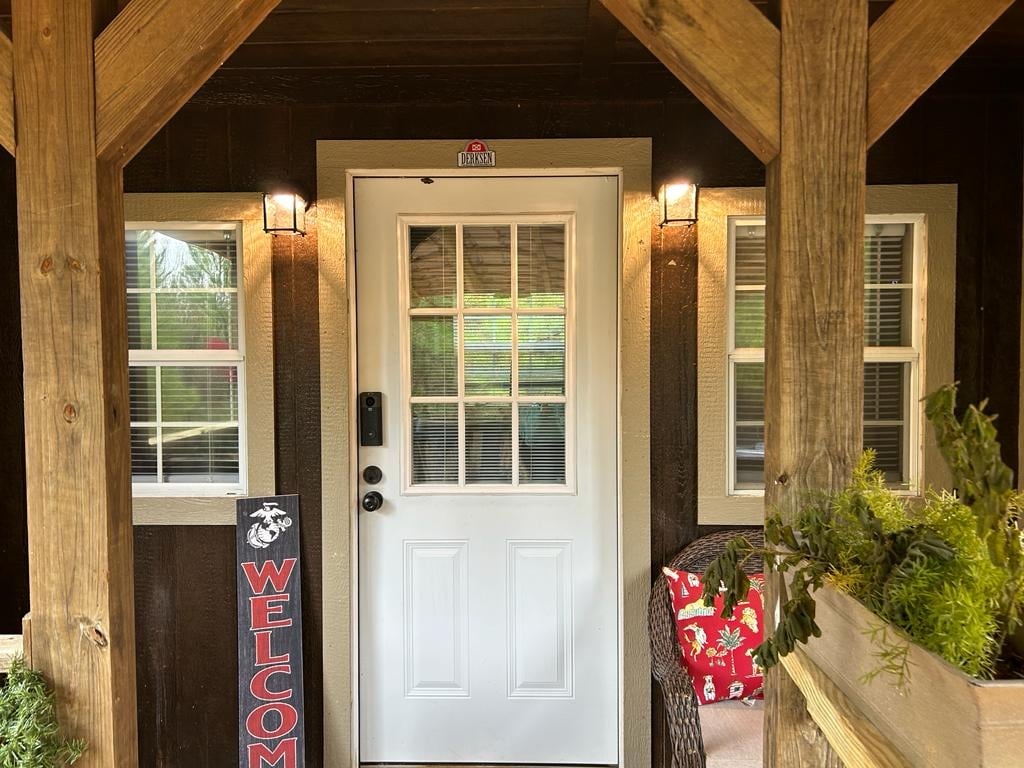 A welcoming front entrance features a white door with a grid of small windows, framed by two adjacent windows. Warm lighting is provided by sconces on either side. A decorative 'WELCOME' sign stands prominently, alongside a pair of chairs adorned with bright cushions.