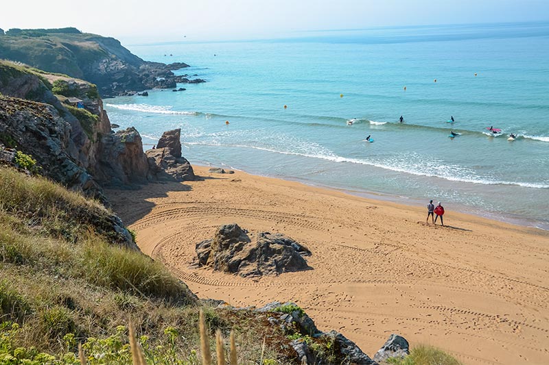 A sandy beach stretches along the coastline, with gentle waves lapping at the shore. Natural rocky formations are visible along the beach, surrounded by grass and low vegetation. Surfers can be seen in the distance, enhancing the serene beach environment.