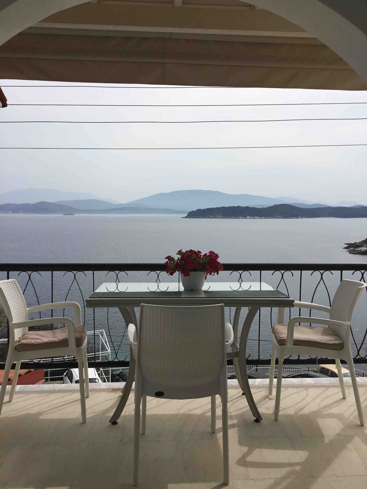 A balcony offers a table with four white chairs, adorned with a flower pot. The view overlooks calm waters and distant islands, framed by soft mountains under a hazy sky.