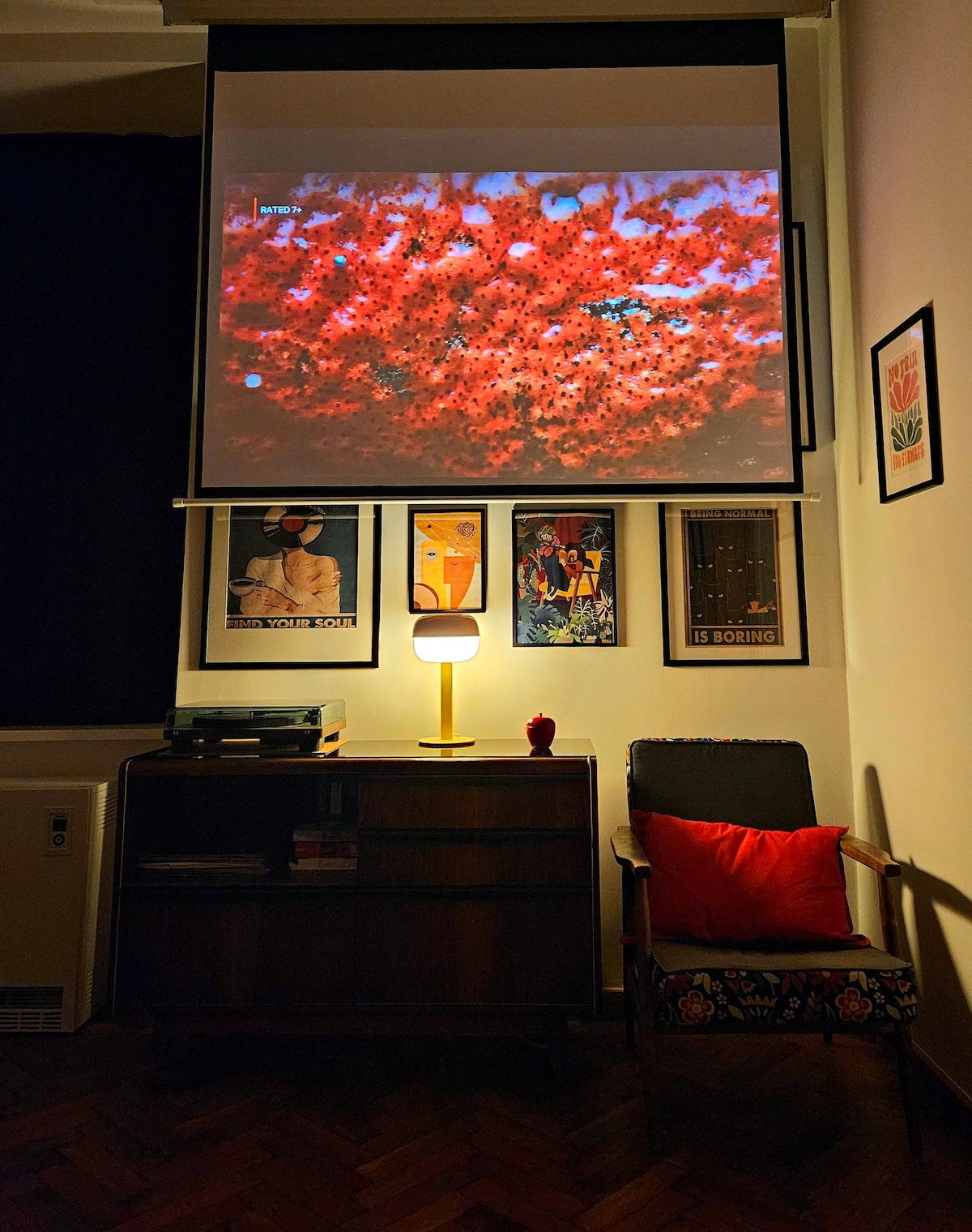 A cozy reading nook features an armchair adorned with colorful fabric and a red cushion. A low wooden cabinet holds a turntable and various records. On the wall, framed artwork adds character, while a projector screen displays vibrant imagery. Soft lighting creates an inviting atmosphere.