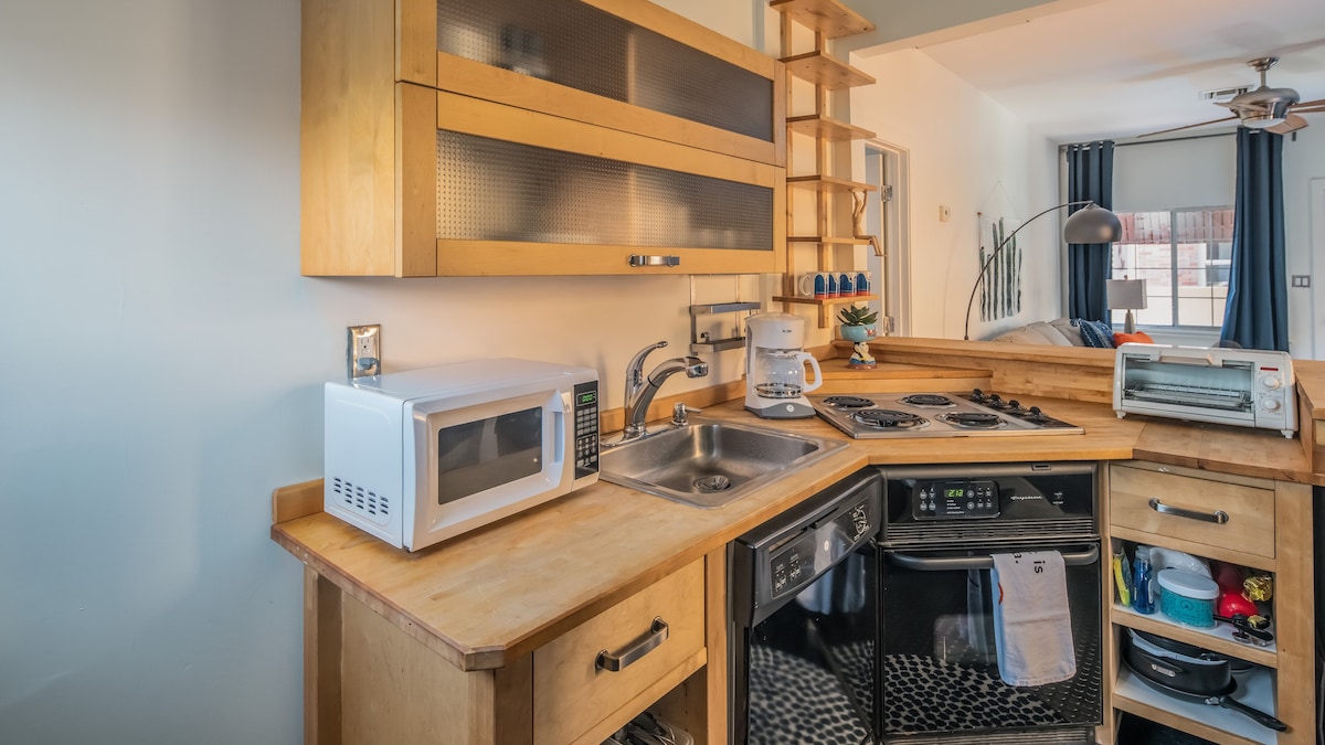 A compact kitchen features warm wooden cabinetry with glass-front storage. Essential appliances, including a microwave and toaster, are present alongside a stainless steel sink and a stove with multiple burners. A neutral, tiled backsplash complements the overall design, creating a functional cooking space.