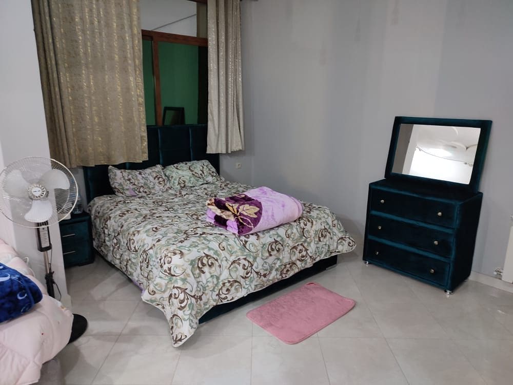 The sleeping area features a spacious bed with an elegant floral comforter. A plush purple blanket is folded at the foot of the bed. Natural light enters through partially draped window treatments, and a stylish dresser with a mirror complements the decor.