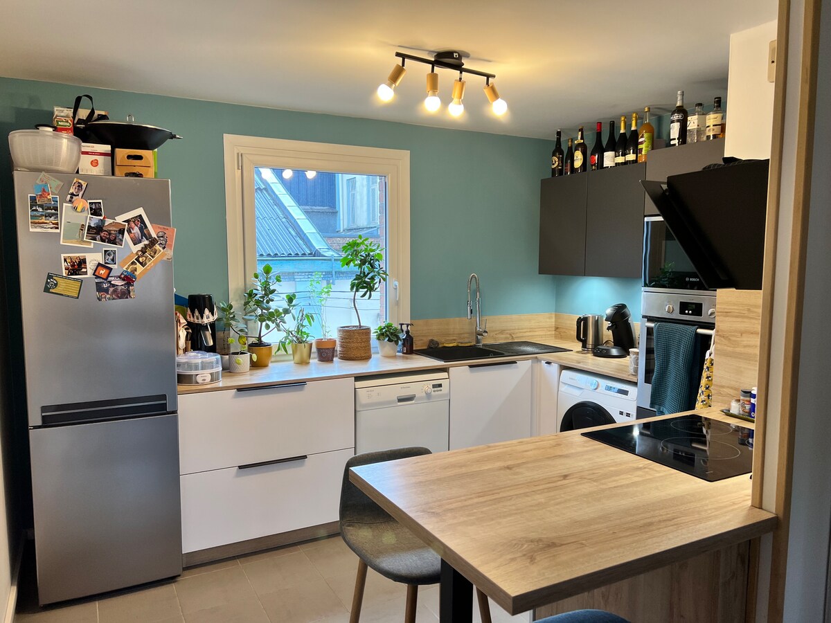 A modern kitchen designed with light wood and white cabinetry, features stainless steel appliances including a refrigerator and dishwasher. A spacious window allows natural light to illuminate the area, while plants and kitchen items add a homey touch. A small dining table is equipped with two stools.