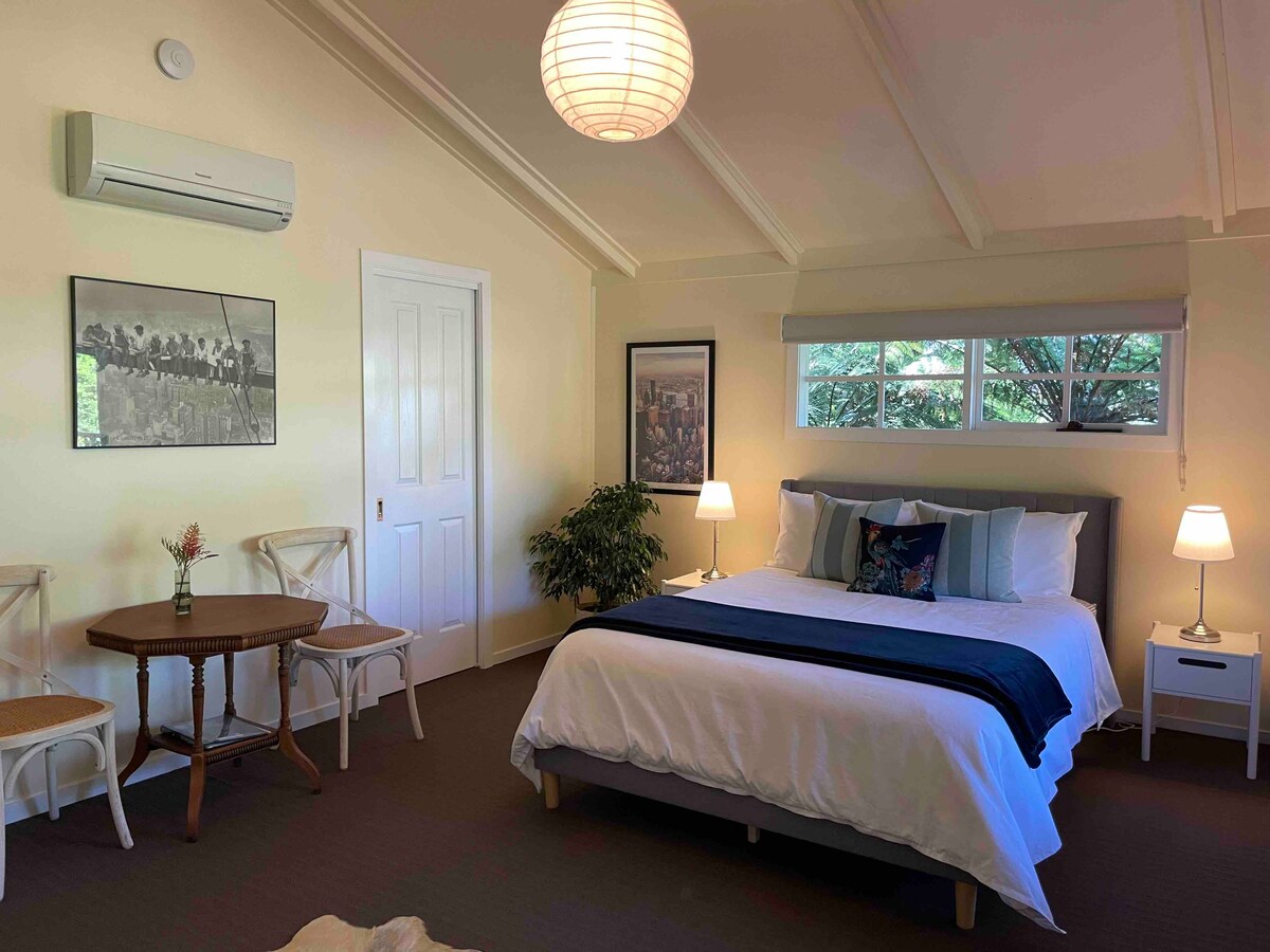 A spacious room is furnished with a queen size bed, accented by decorative pillows. Nearby, a small dining table with chairs is positioned beside a plant. Two bedside lamps illuminate the space, complemented by natural light from the windows above the bed.