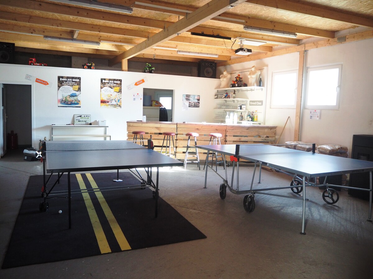 An open communal space is showcased, featuring several movable tables on wheels and a minimalist kitchen area in the background. A bar with seating is positioned against the wall, and light enters from numerous windows, creating a bright atmosphere.