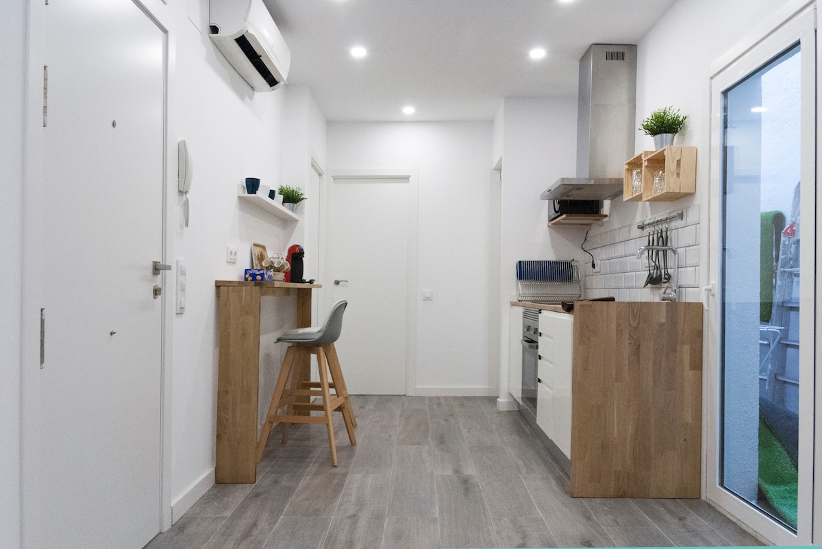 A bright kitchen area is seen, featuring modern cabinetry and stainless-steel appliances. A wooden countertop with two stools provides space for dining. Natural light enters through a sliding glass door, which leads to an outdoor area. The walls are painted in a neutral tone.