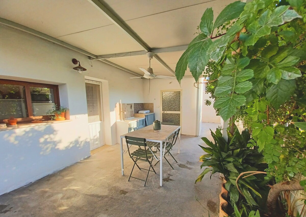 An outdoor dining area is set beneath a covered patio, featuring a long table with folding chairs. Lush greenery surrounds the space, while natural light filters through the open doorway, enhancing the connection between indoor and outdoor spaces.
