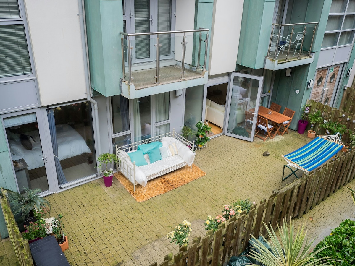 A spacious outdoor patio is featured, surrounded by greenery. Comfortable seating, including a white couch with turquoise cushions, is highlighted. Tables and a hammock are arranged for relaxation. The patio is accessible from the adjacent living area through large glass doors.