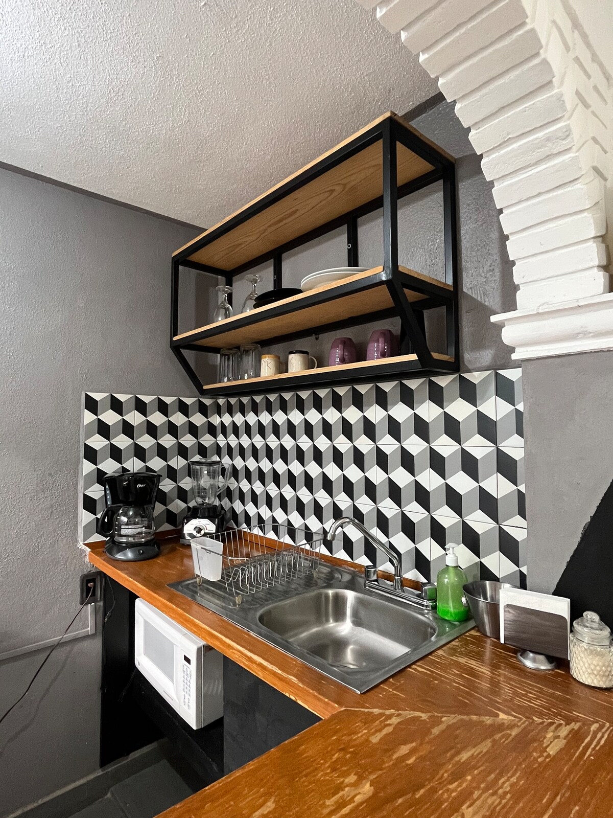 A modern kitchen space showcases geometric patterned tiles in black, white, and grey. A wooden countertop complements a stainless steel sink and various appliances, including a coffee maker and a microwave. Open shelving displays glassware and decor, enhancing the functionality of the area.