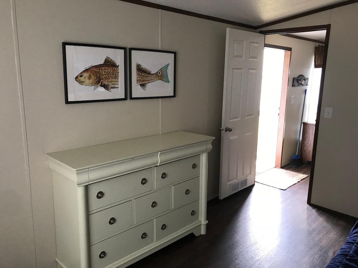 A light-colored dresser with six drawers is positioned against a wall, adorned with two framed fish prints. The open door in the background reveals a glimpse of a small entryway, enhancing the room's openness and accessibility.