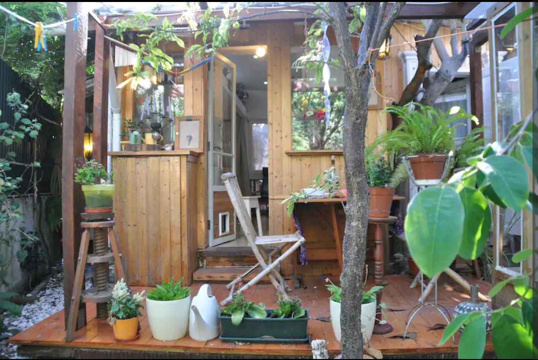 A cozy wooden deck features various potted plants and a small chair, surrounded by greenery. The entrance is framed by open doors, inviting natural light into the interior. A wooden table stands against the wall, enhancing the serene atmosphere.