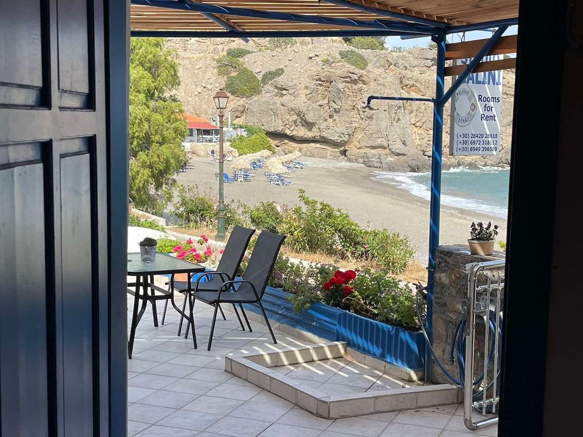 A courtyard area is visible, featuring a dining table with chairs and vibrant flowers along the edge. Beyond, the sandy beach and calm sea are seen, framed by natural rock formations. Coastal lounge chairs are arranged along the shoreline, with a clear sky above.