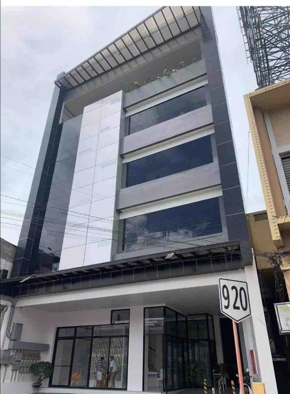 A modern, five-story building is depicted, featuring sleek glass and white paneling. The ground floor showcases large windows, allowing for ample natural light. The roof is adorned with greenery, enhancing the contemporary design of the structure.