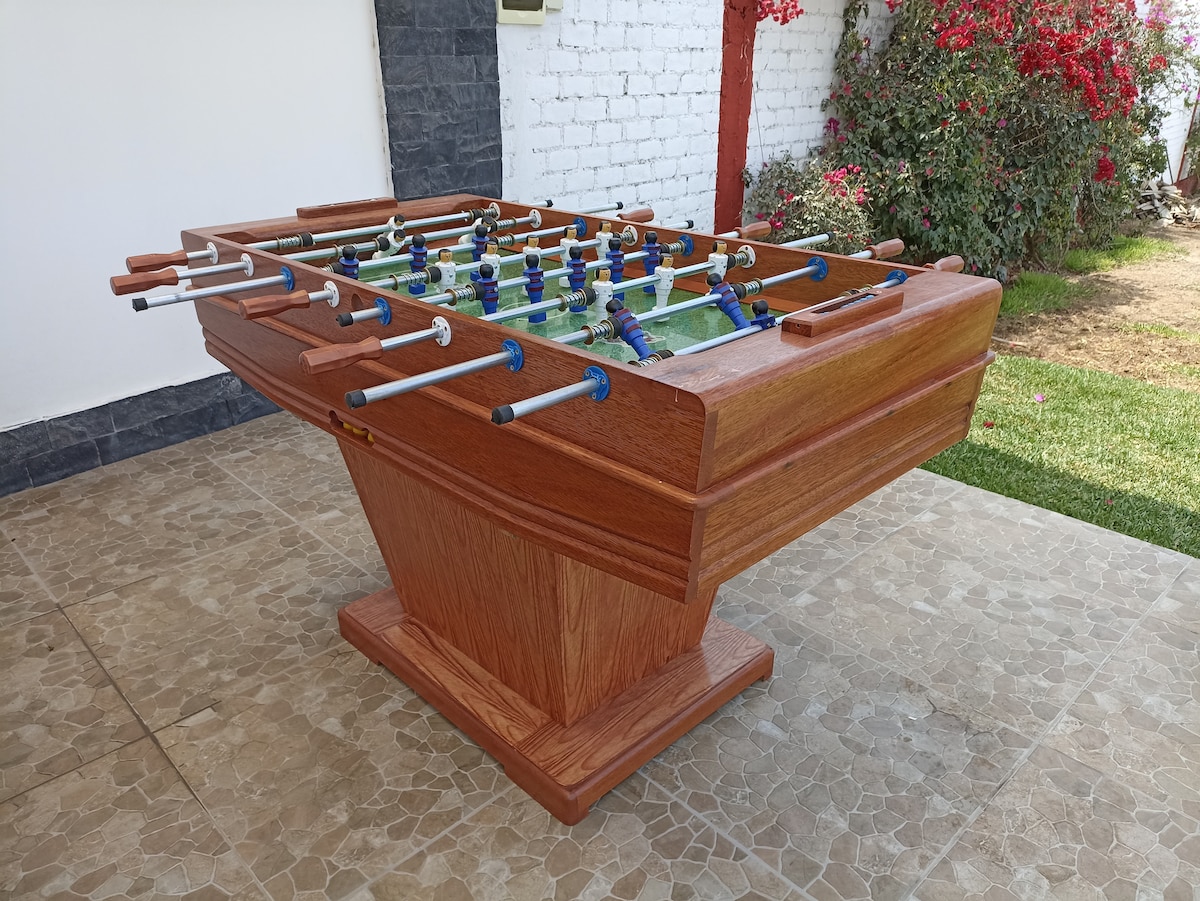 A wooden foosball table is displayed outdoors in a well-maintained area. The table features multiple player rods and a vibrant green playing field. Surrounding the table is a grassy space, complemented by colorful flowering plants in the background.