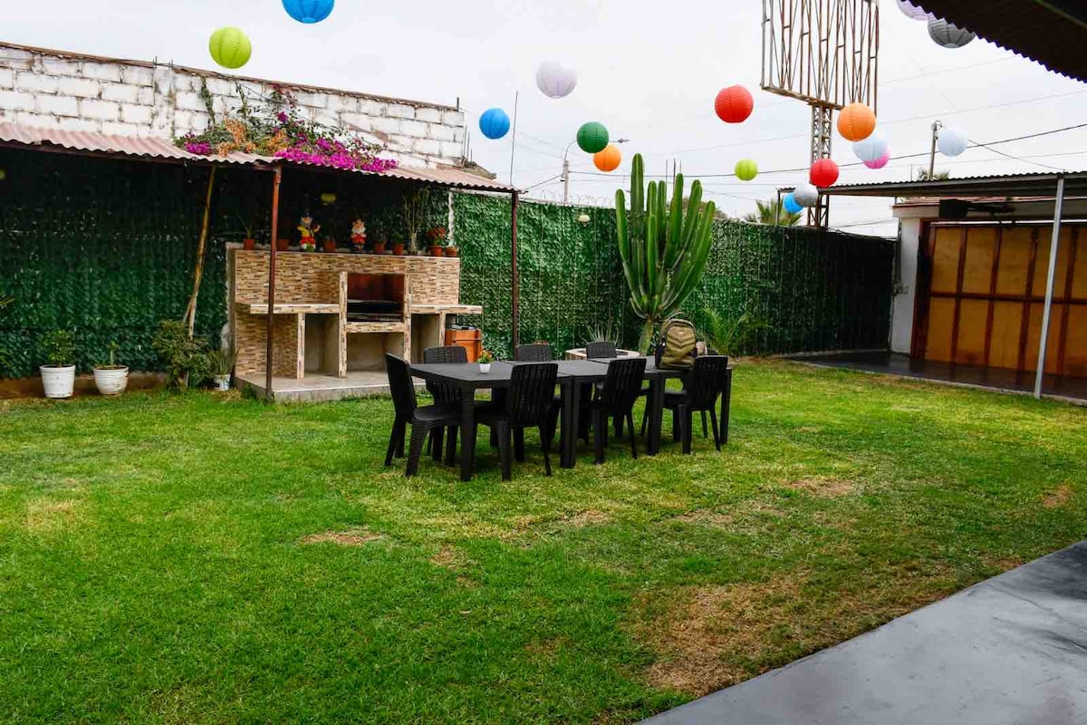 An outdoor space features a manicured grassy area bordered by vibrant green plants and colorful hanging lanterns. A decorative barbecue area is present alongside a large table surrounded by black chairs, creating an inviting setting for gatherings.