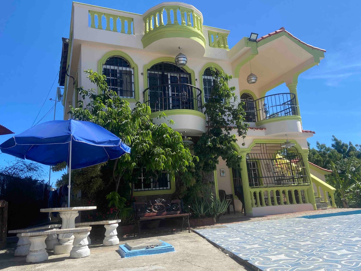 A three-story building features a bright exterior with green accents and ample windows. A shaded seating area with a large umbrella is visible in the foreground, accompanied by a concrete table. Lush greenery surrounds the property, creating a connection with nature.
