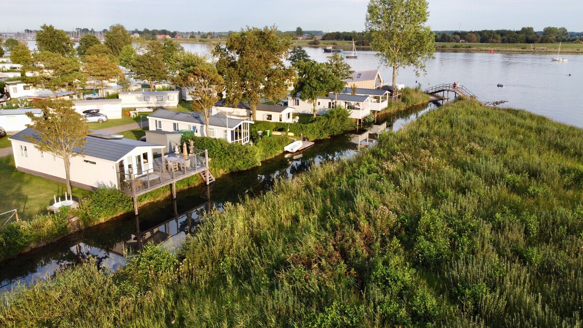 An aerial view captures a cluster of modern lodges set along a tranquil waterway, surrounded by lush greenery. The gently flowing water reflects the sky, while neighboring trees provide a natural border, enhancing the serene atmosphere of the vacation park.