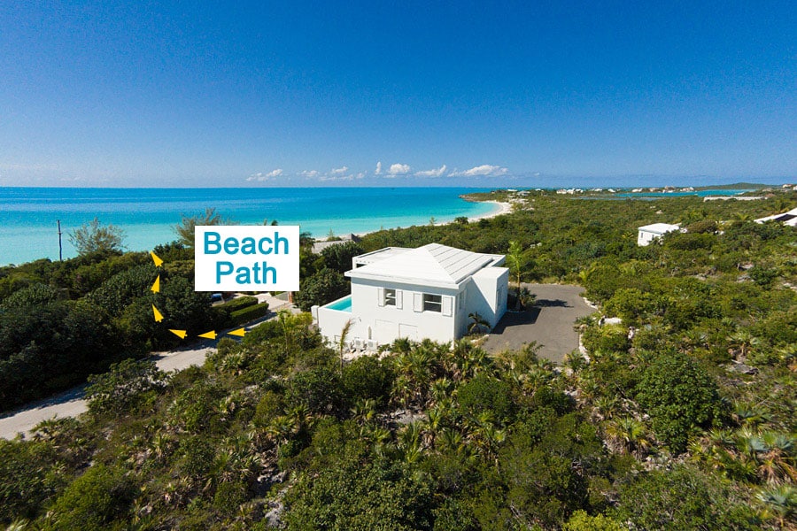An aerial view of the villa is presented, showcasing its proximity to the tranquil beach. A clear path leading to the beach is marked, surrounded by lush greenery and panoramic views of the turquoise ocean and sandy shore.
