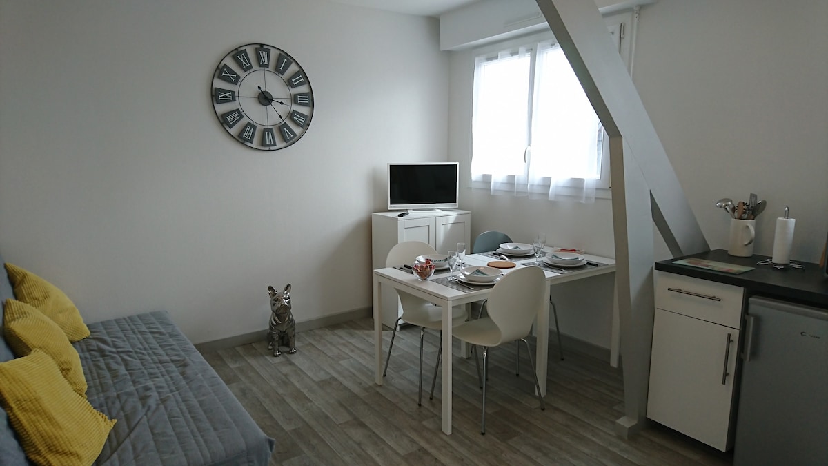 A bright living area features a cozy sofa adorned with yellow cushions. A dining table set for four is positioned nearby, complemented by light-colored chairs. A clock adorns the wall, and a television is visible on a cabinet, with large windows providing ample natural light.