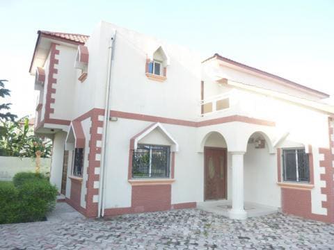 A two-story villa is displayed, featuring a combination of brick and smooth white walls. Arched entryways and window frames add architectural interest. The landscaped exterior includes neatly trimmed greenery and a paved path leading to the entrance.