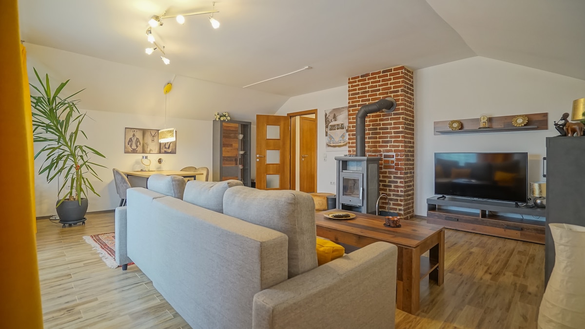 A spacious living area is depicted, featuring a large gray sofa and a wooden coffee table. A brick fireplace serves as a focal point, while a flat-screen TV is placed on a dark wood cabinet. Natural light illuminates the room, complemented by decorative plants.
