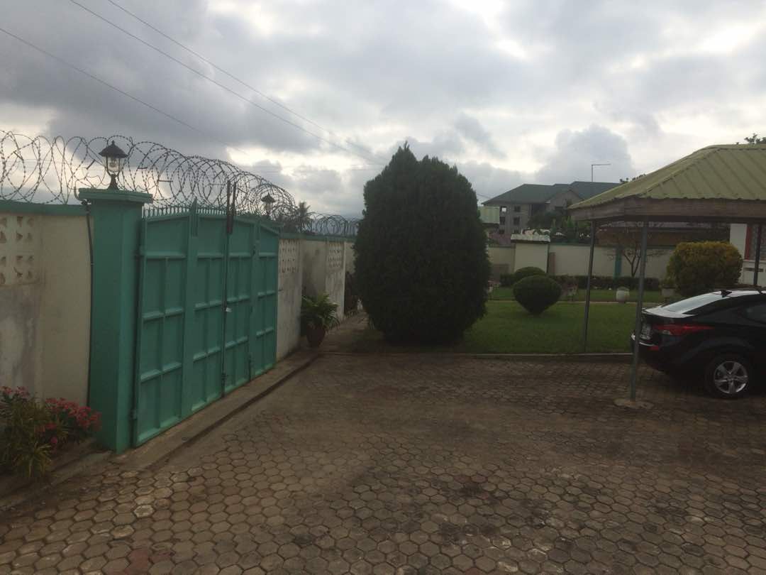 A secured entrance is depicted, featuring a turquoise gate with barbed wire atop. The driveway is paved with stones, bordered by greenery, including a neatly trimmed shrub and a parked black car. The surroundings are well-maintained, contributing to a tranquil atmosphere.