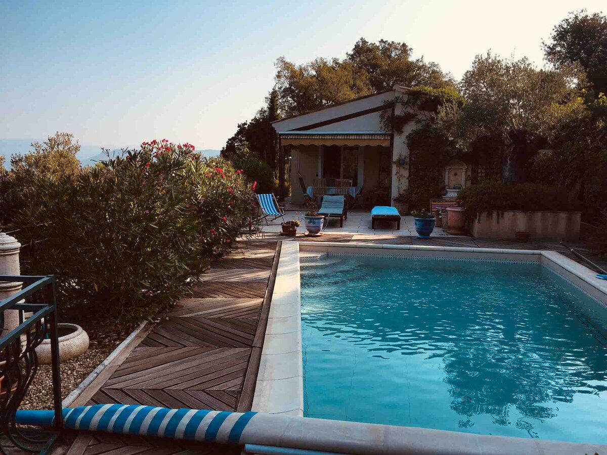 A serene pool area is framed by lush greenery and flowering plants. The inviting swimming pool, surrounded by wooden decking, reflects the sunlight. Comfortable loungers are positioned nearby, with a shaded seating area visible in the background, providing a tranquil setting for relaxation.