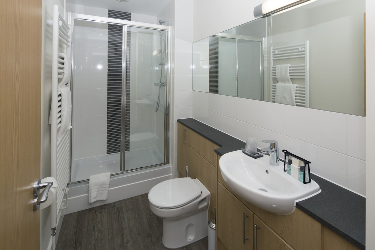 A modern bathroom features a glass shower enclosure and a sleek toilet. A spacious countertop with a sink is complemented by wooden cabinetry. A mirror spans the wall above the sink, enhancing the room’s brightness, while towel racks provide additional convenience.