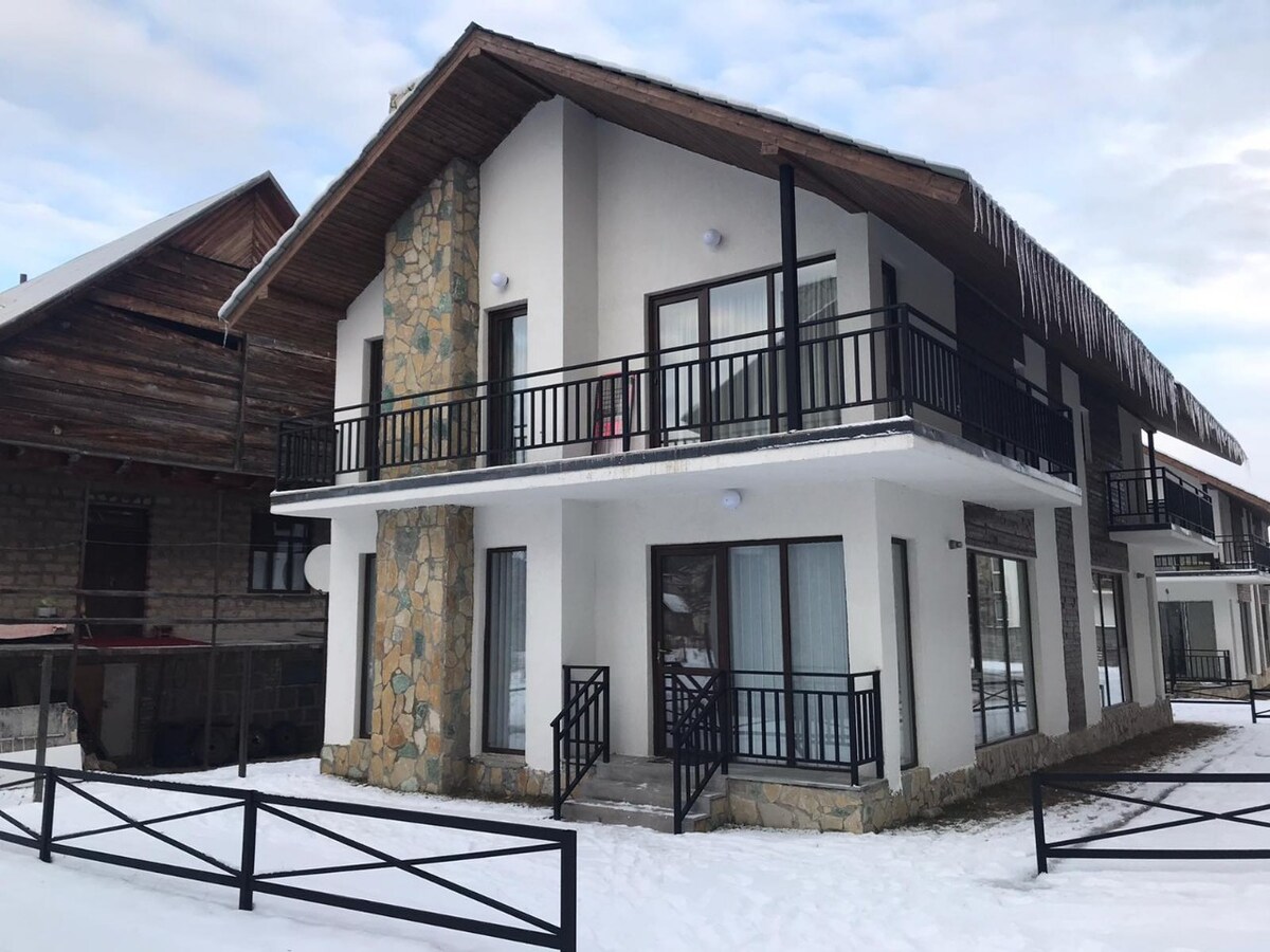 A two-story building is shown, featuring a blend of modern and rustic design elements. The exterior is characterized by white walls, a stone-clad section, and large windows. Snow covers the ground, enhancing the winter setting.