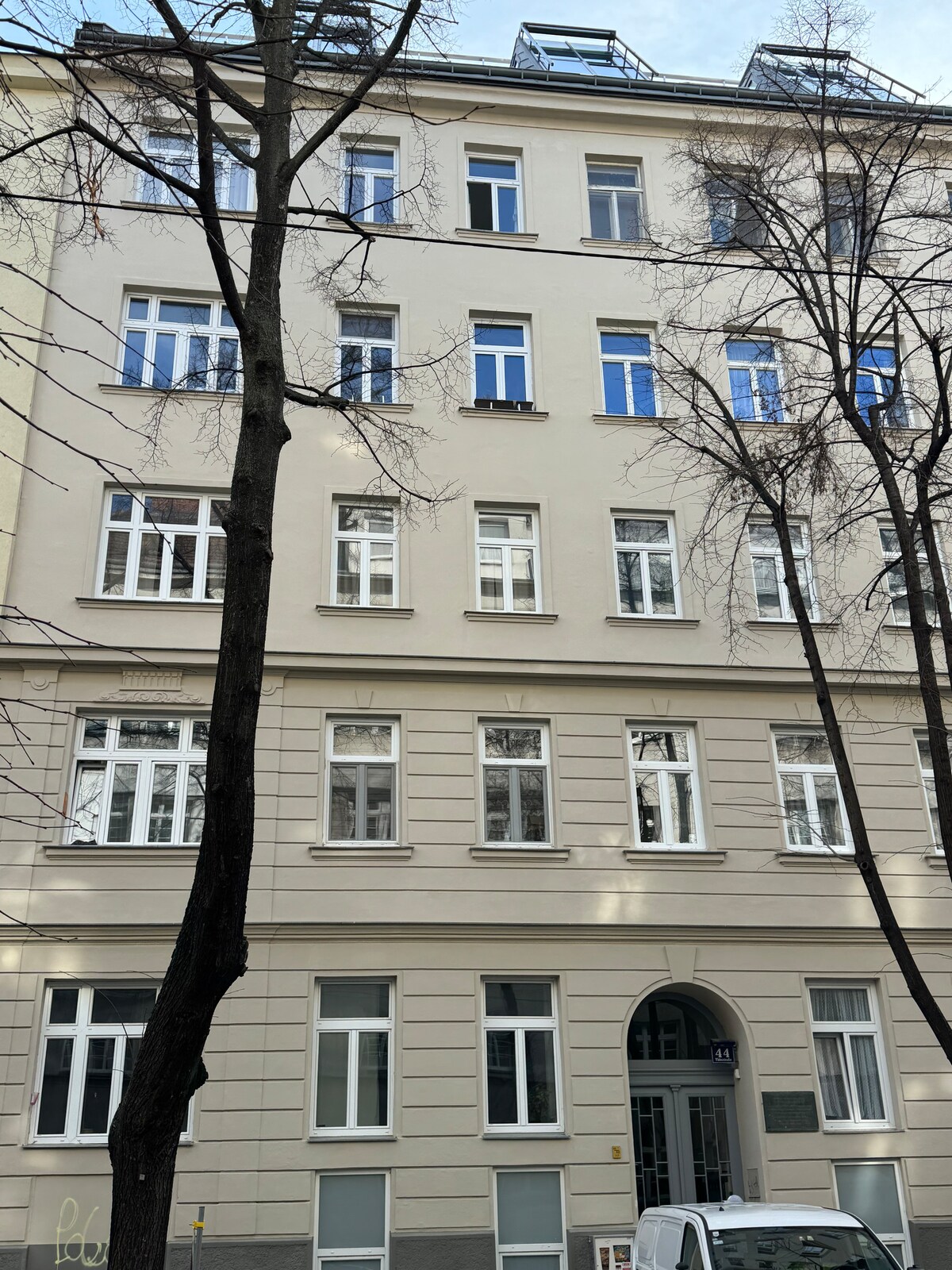 A multi-story building is shown from the street, featuring a classic facade with a combination of large windows and smaller units. The entrance is framed by symmetrical architectural details, while trees line the sidewalk, providing a natural element to the urban scene.