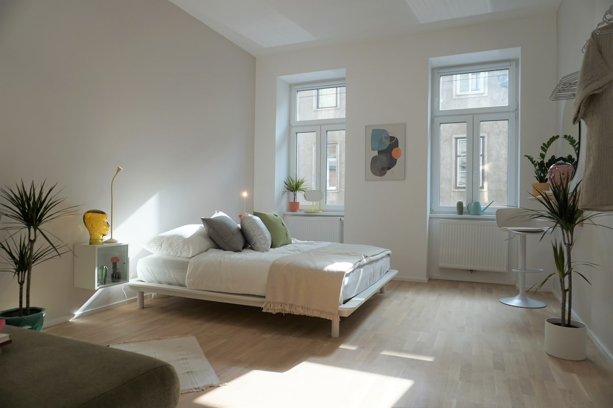 A spacious sleeping area is illuminated by natural light from two large windows. A double bed is centered in the room, with a light blanket draped casually. Decorative plants and modern artwork in soft tones add a touch of elegance to the minimalist design.