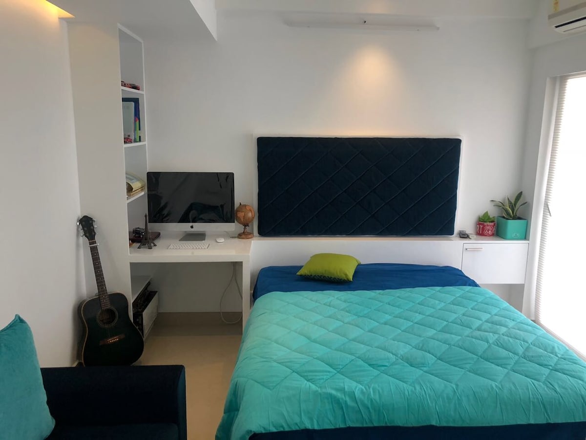 A serene sleeping area features a large bed dressed in a turquoise blanket, complemented by a contrasting green accent pillow. A dedicated workspace with a computer is arranged beside the bed, while a cozy armchair and a decorative plant add a touch of comfort and style.