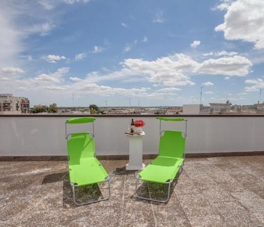 An outdoor terrace features two lounge chairs in bright green positioned beside a small table. The patio provides an open view of the sky with scattered clouds. Urban scenery and rooftops can be seen in the background.