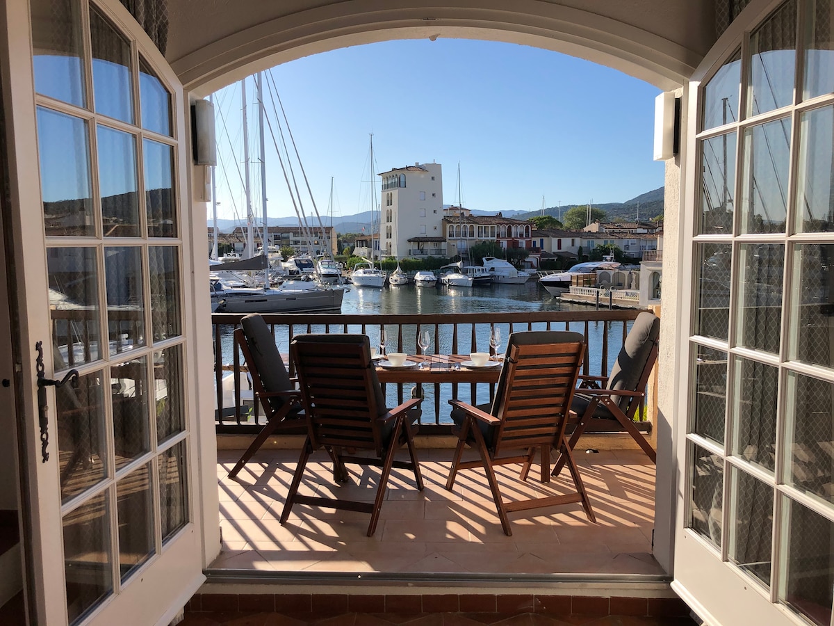 A set of wooden reclining chairs and a small table is arranged on the private terrace, facing the marina. Panoramic views of boats and waterfront buildings are visible, with clear blue skies and gentle sunlight enhancing the tranquil scene.