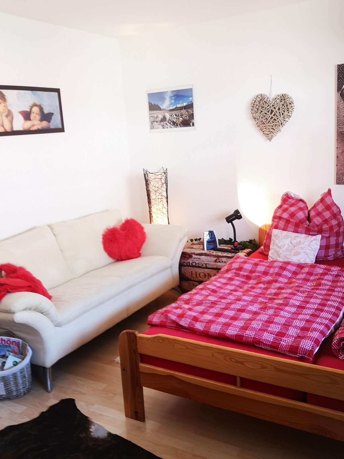 A cozy room features a spacious bed adorned with a red checkered blanket, accompanied by a white sofa with decorative pillows. A standing lamp and a small desk lamp provide additional lighting. Wall art adds character, while natural light enhances the inviting atmosphere.