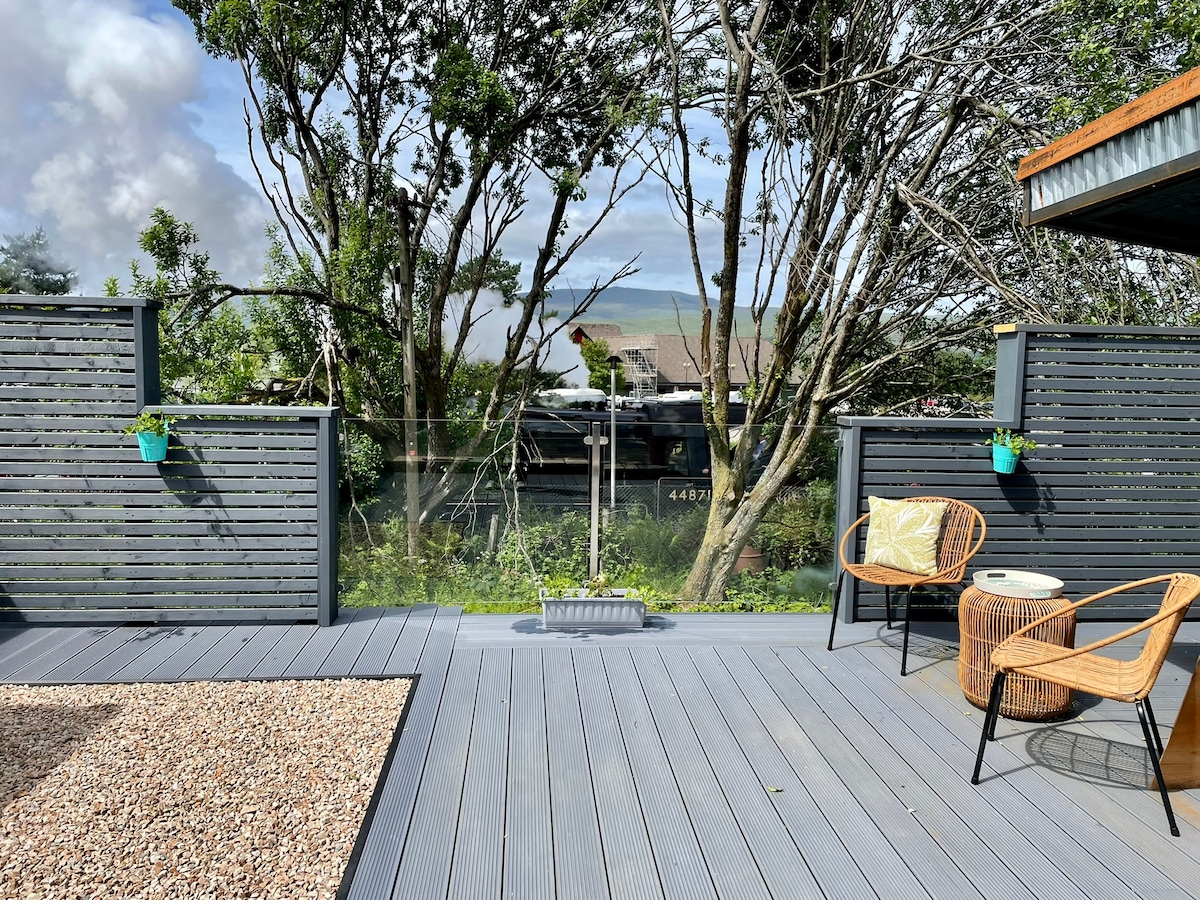A large deck features a mix of gravel and composite flooring, surrounded by wooden privacy screens. Two stylish chairs are positioned for outdoor relaxation, with small potted plants adding a touch of greenery. The view showcases distant trees and the landscape, creating a peaceful setting.